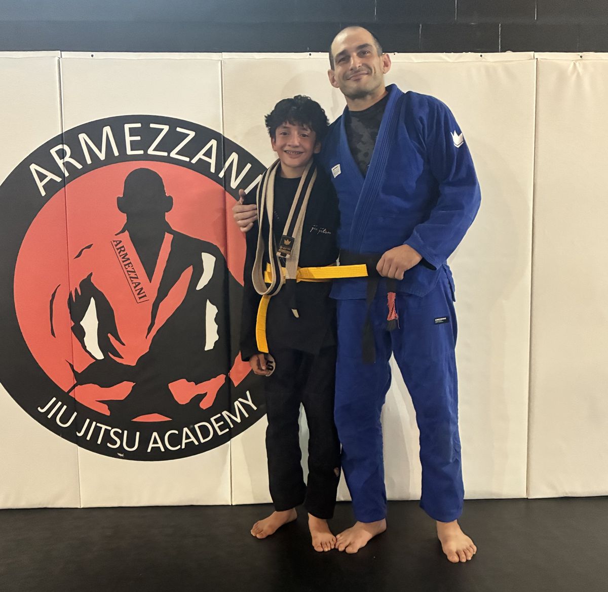 Smiling kids student with his new yellow belt next to Professor Lou in front of the academy logo